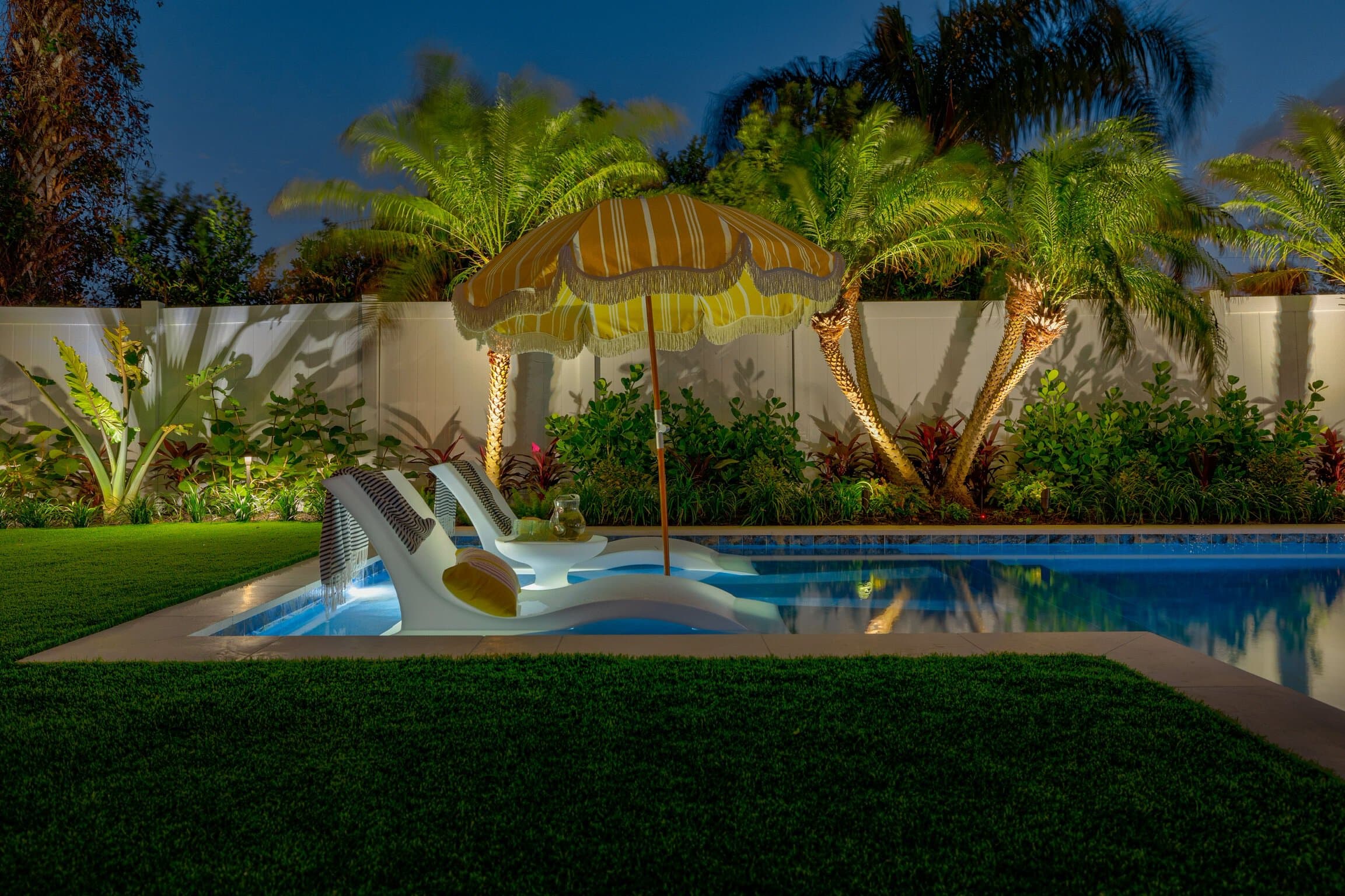 Night view of backyard pool with lounge chair, umbrella, and tropical plants.